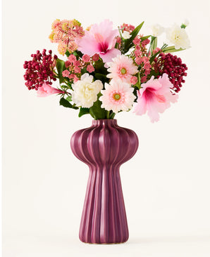 Een boeket kunstbloemen van Zijden, ‘Bloom & Berry’, met roze, witte en rode kunstbloemen, is prachtig geschikt in een hoge, geribbelde, paarse vaas tegen een effen witte achtergrond.