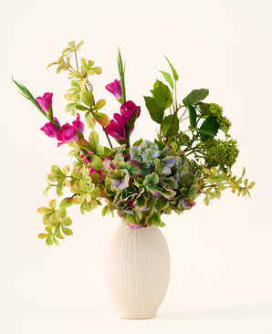 In een witte vaas met structuur staat een boeket van groene hortensia’s, bladrijke takken en felmagenta zijden bloemen tegen een effen lichte achtergrond, waardoor een opvallend ‘Cottage Garden’-boeket van zijden kunstbloemen ontstaat dat elke ruimte een blijvende charme geeft.