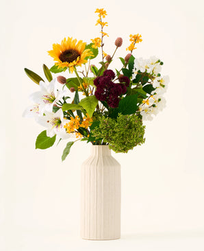 In een crèmekleurige vaas staat het Zijden kunstbloemenboeket ‘Mom’s Garden’, een arrangement van zonnebloemen, lelies, witte en bordeauxrode bloemen en groen blad tegen een effen achtergrond — waardoor het eruitziet als een kunstbloemenboeket dat doet denken aan een zomerse tuinborder.