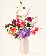In een witte keramische vaas staat het boeket ‘Rosy Revery’ van Zijden kunstbloemen, een levendig arrangement van roze, paarse, oranje en rode bloemen met groene bladeren en stelen, tegen een lichte achtergrond.