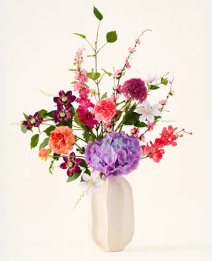 In een witte keramische vaas staat het boeket ‘Rosy Revery’ van Zijden kunstbloemen, een levendig arrangement van roze, paarse, oranje en rode bloemen met groene bladeren en stelen, tegen een lichte achtergrond.