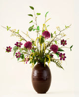 In een bruine, geribbelde vaas staat het boeket „Woodland Charm“ van Zijden kunstbloemen, met paarse alliums, donkerroze bloemen, gele takjes en groen blad tegen een effen, lichte achtergrond.
