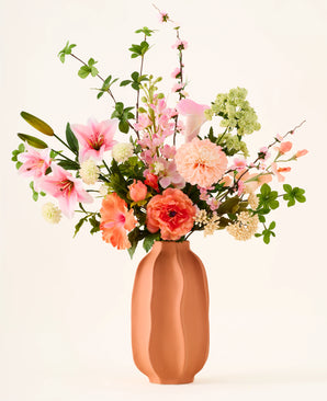 In een terracotta vaas staat het kleurrijke boeket ‘Yasmine’s Choice’ van zijden kunstbloemen, bestaande uit roze lelies, perzikkleurige rozen en groen blad, vervaardigd uit hoogwaardige zijden bloemen van Easyplants, allemaal prachtig gearrangeerd tegen een lichte achtergrond.