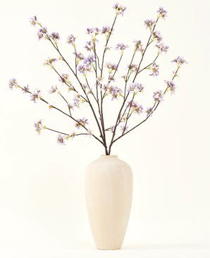 Een hoge, crèmekleurige vaas van keramiek bevat een aantal dunne takken met de tekst Zijden kunstbloemen boeket Kunstbloementak paars, tegen een effen, lichte achtergrond.