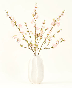 Het Zijden kunstbloemen boeket Kersenbloesem lichtroze 94 cm heeft delicate roze kunstmatige kersenbloesems in een witte keramieken vaas, die elegantie toevoegt tegen een effen lichte achtergrond.