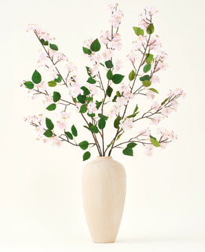 Een beige vaas van keramiek bevat verschillende lange takken met het Zijden kunstbloemen boeket Perzikbloesem roze en groene bladeren, tegen een effen witte achtergrond - perfect voor een kunstbloemen interieur.