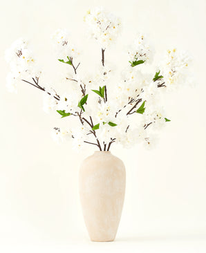 Het Zijden kunstbloemen boeket Kersenbloesem crème 119 cm heeft elegante bruine takken met zacht witte nepbloesem en groene bladeren, weergegeven in een eenvoudige beige vaas tegen een effen lichte achtergrond.